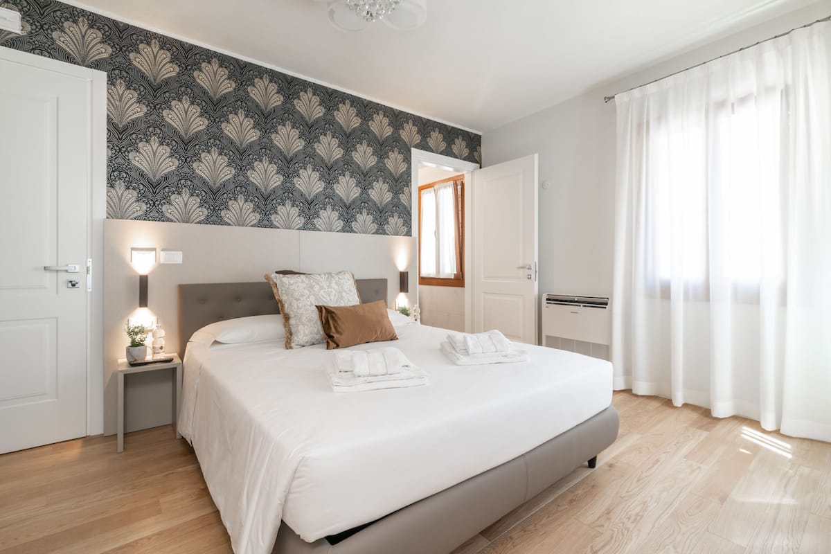 A bedroom with a large bed, two bedside tables, a nightstand, a wall-mounted light, and a window with curtains.
