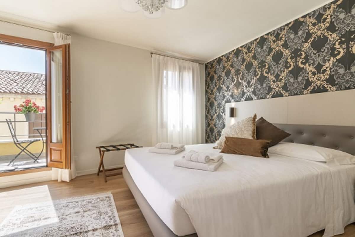 A bedroom with a large bed, a nightstand, a window with curtains, and a view of the outside balcony.