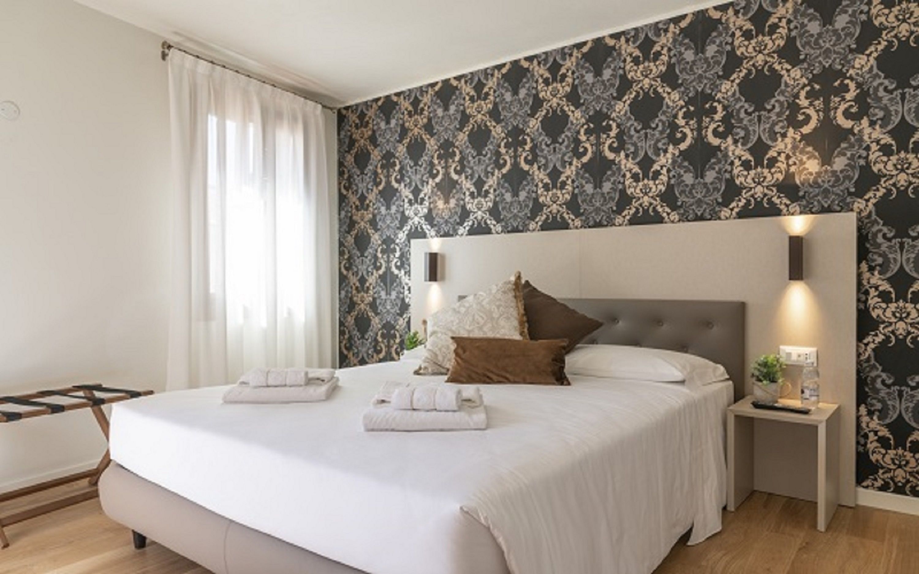 A neatly made bed with white linens and two towels, a bedside table with a lamp, and a wall with a patterned wallpaper.