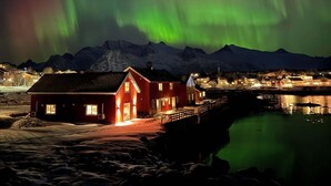 A red building with a dock extending into a body of water, surrounded by snow-covered mountains under a night sky with green aurora. - Astra Village - Waterfront Rorbu Cabins in Lofoten (Vagan)