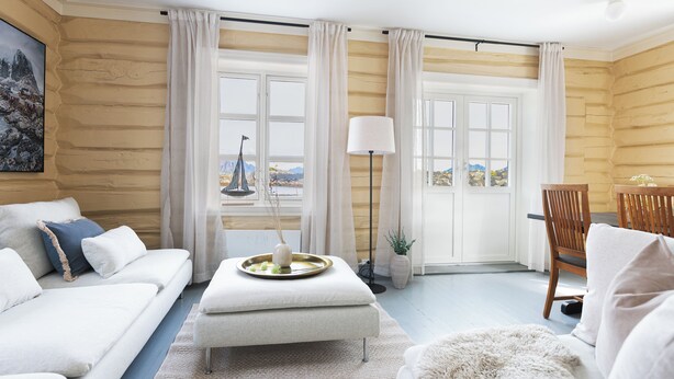 A log cabin interior with a white sofa, a dining table with a lamp, and a view of mountains through the windows. - Astra Village - Waterfront Rorbu Cabins in Lofoten (Vagan)