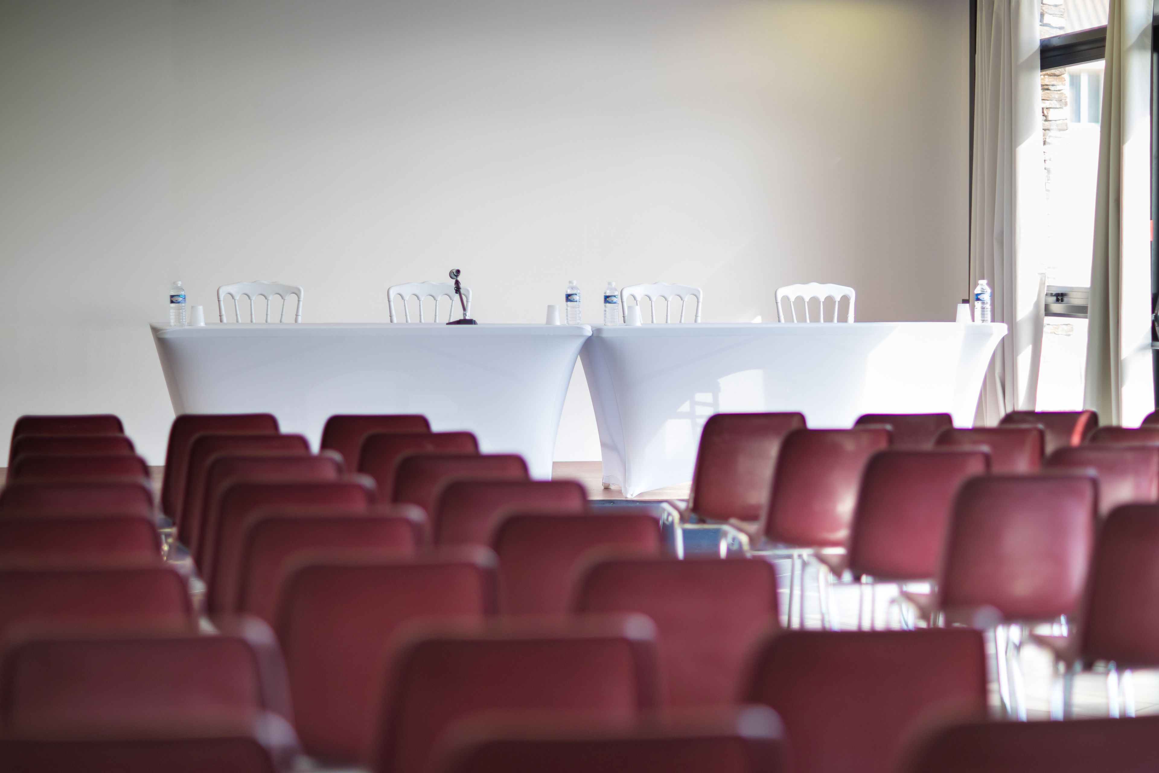 A conference room with rows of red chairs and a white table with water bottles and a microphone.
