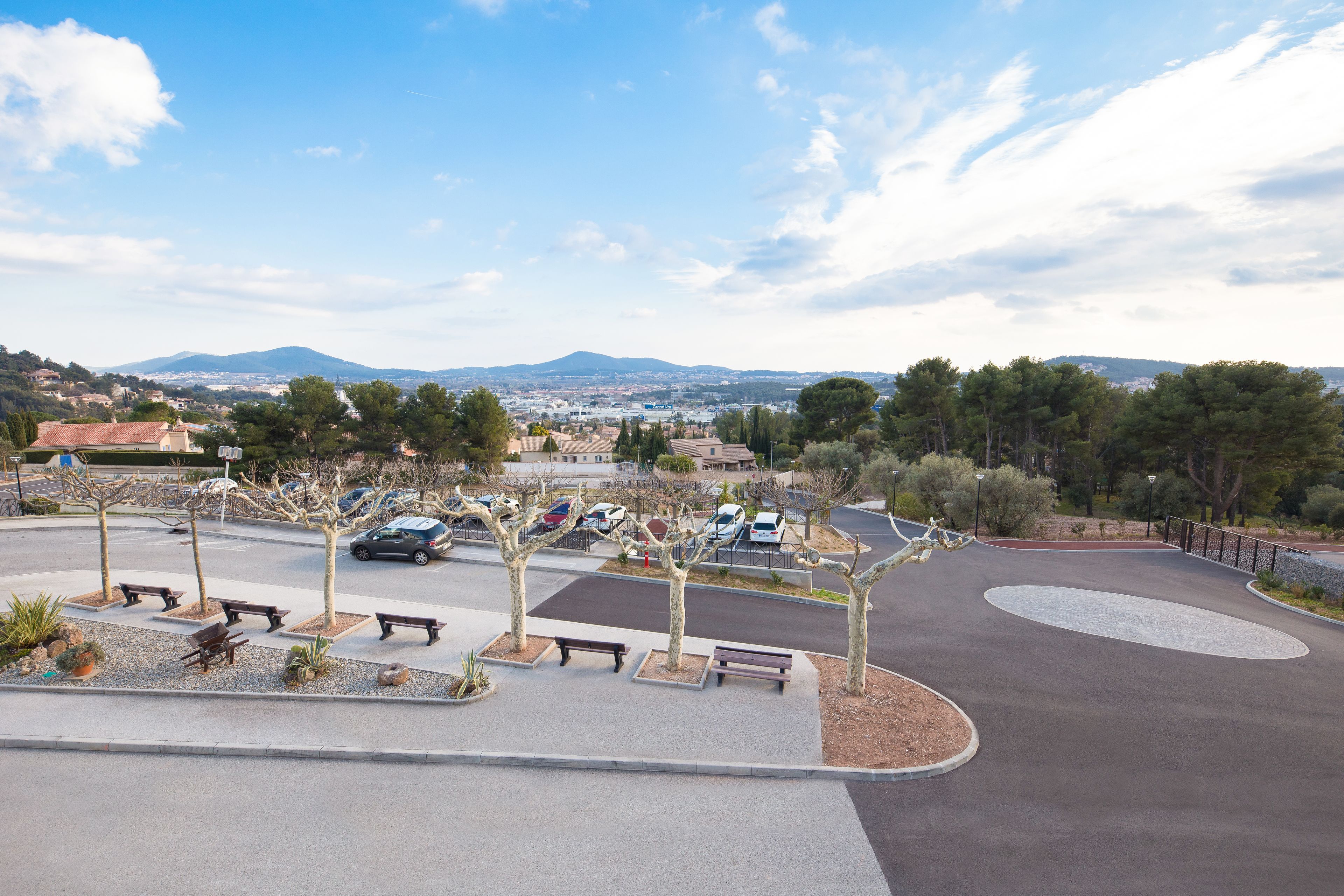 A parking lot with several benches and trees, a curved road, and distant mountains.