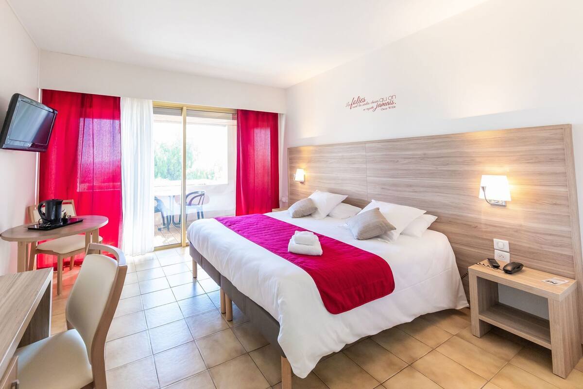 Ein Hotelzimmer mit Bett, Schreibtisch mit Stuhl, Fernseher und einem fensterbepflanzten Fenster.