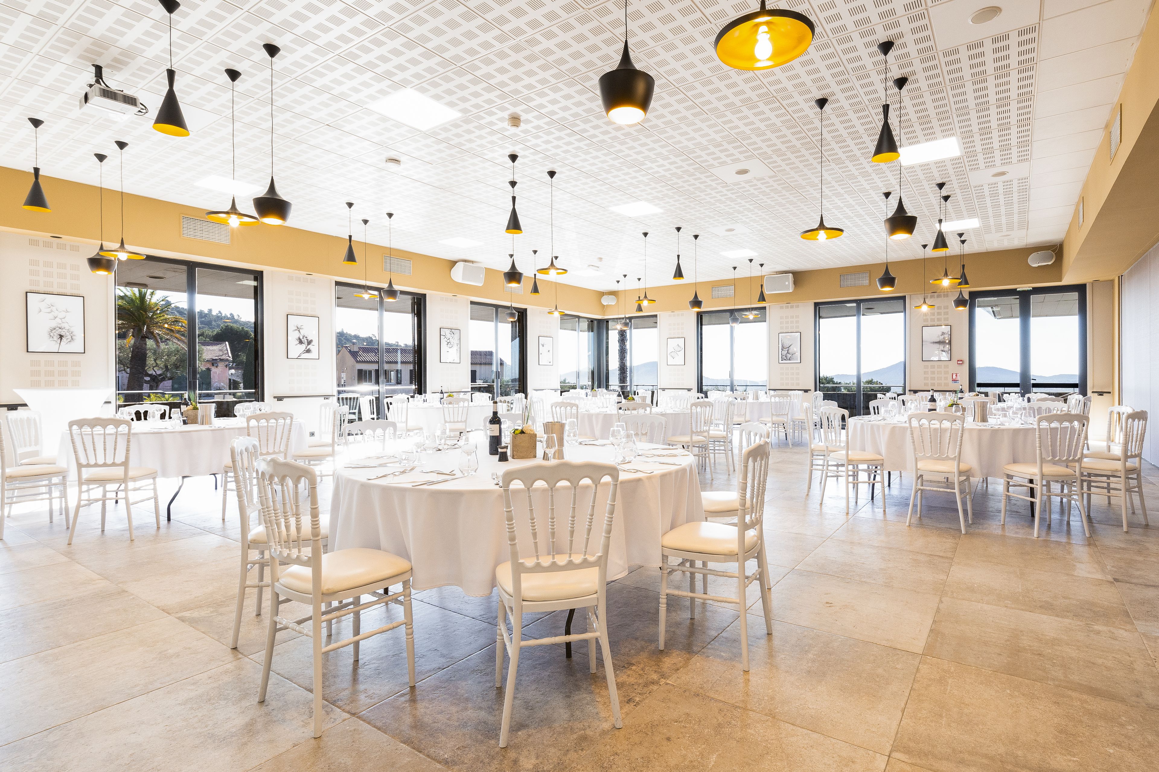 Une salle de conférence spacieuse, dotée de tables rondes, de chaises blanches et d’un éclairage suspendu.