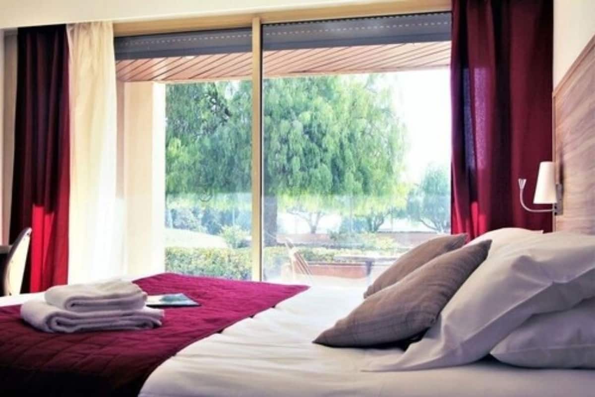 Ein Hotelzimmer mit Bett, einem fenster mit Vorhängen und Blick auf Grünfläche.