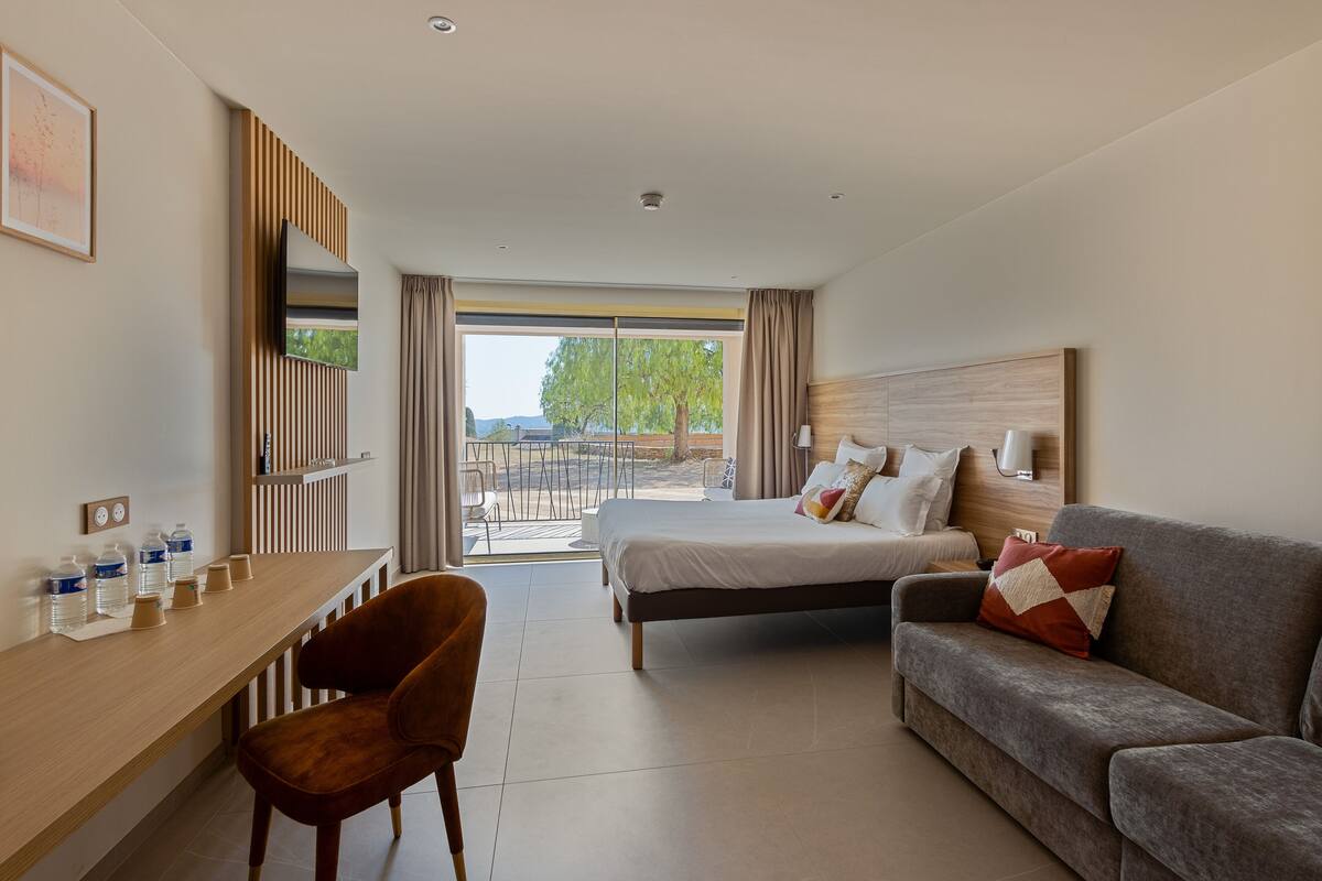 Ein modernes Hotelzimmer mit Bett, einer Couch, einem Schreibtisch und einem Sessel. Ein großes Fenster bietet Blick auf Bäume und ein Gebäude.
