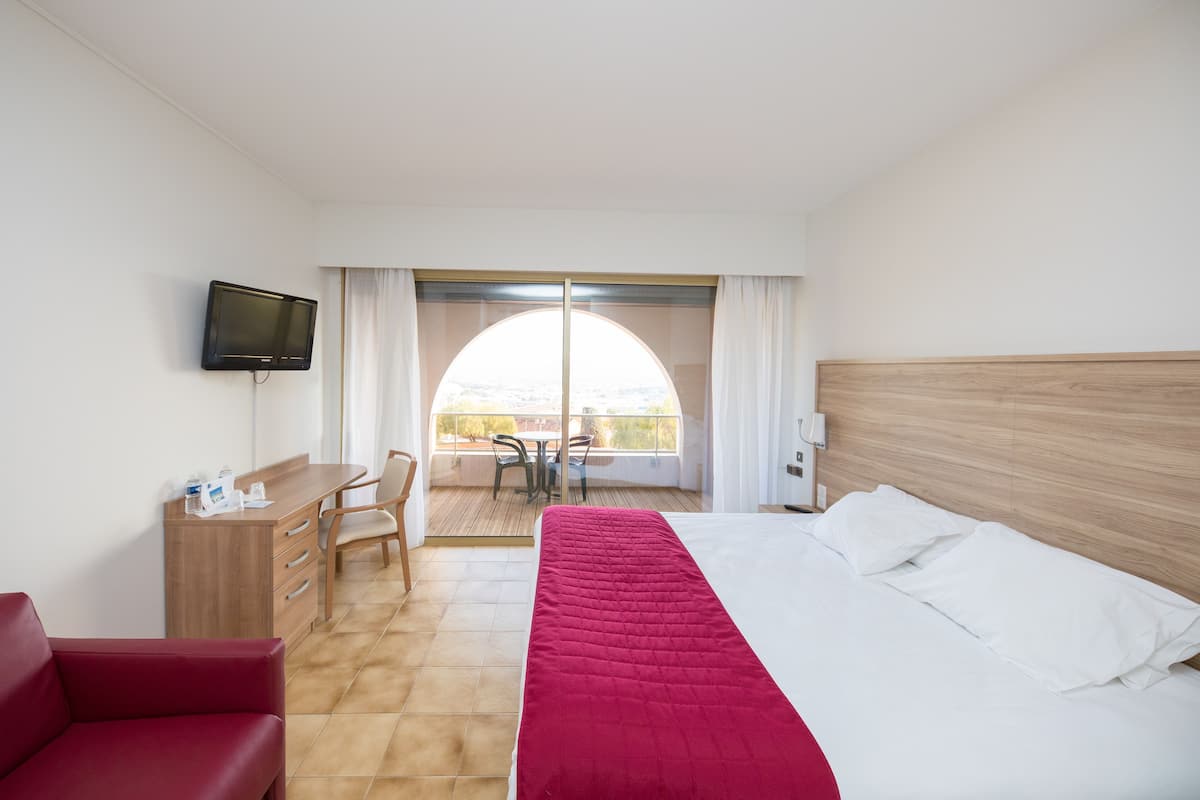 Ein Hotelzimmer mit Bett, Schreibtisch, Fernseher, einem Balkon mit Tisch und Stühlen sowie einem roten Sessel.