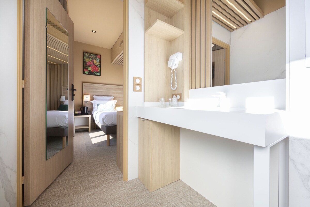 Een moderne hotelkamer met een grote spiegel, een witte wastafel en een bed met witte lakens, en een schilderij aan de muur.