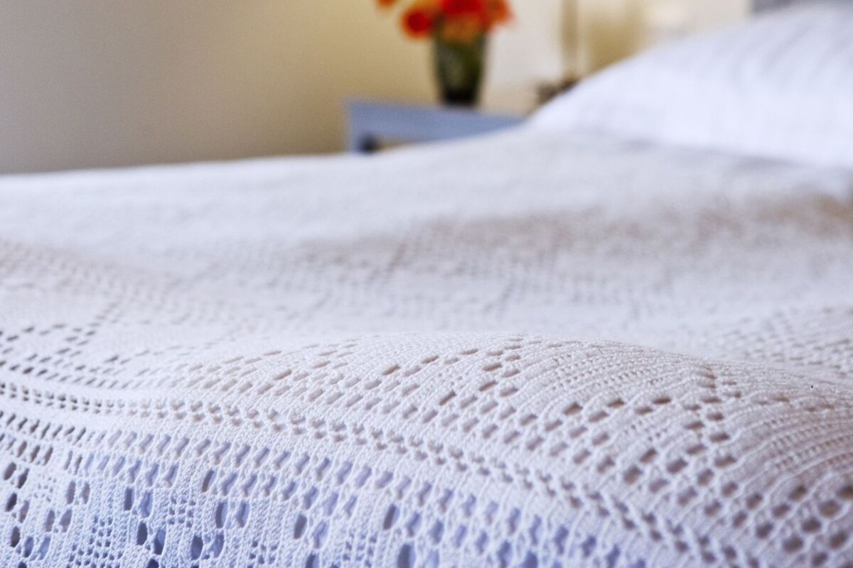 Una cama con una manta blanca y texturizada. En el fondo, hay una lámpara y un florero con flores sobre una mesita de noche.