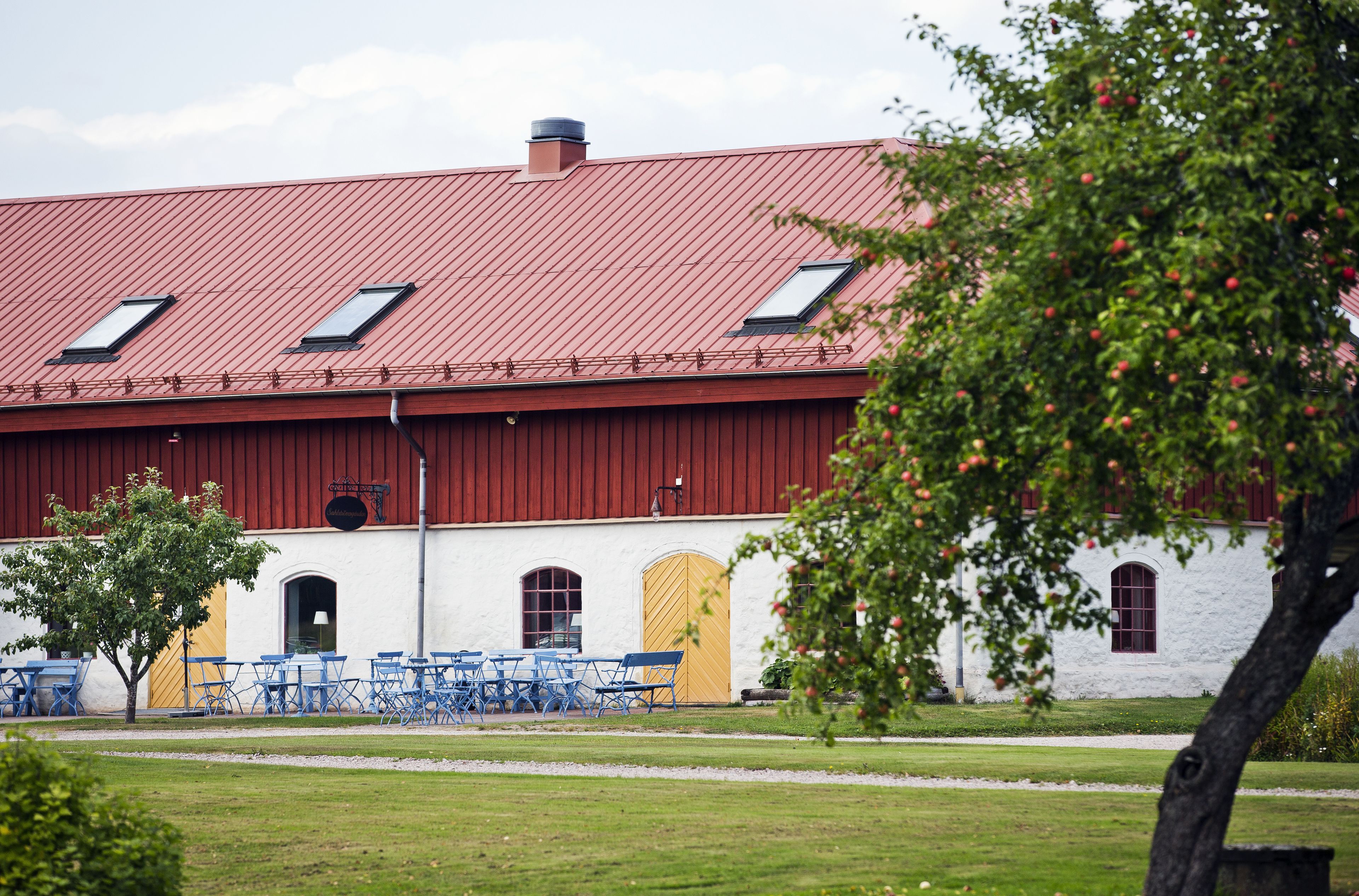 A red-roofed barn with a white exterior, a tree in the foreground, and blue folding chairs outside.
