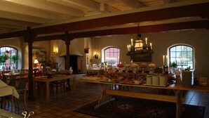 Un comedor espacioso con techos altos, vigas de madera y ventanas amplias. Las mesas están preparadas para una comida, y hay velas sobre ellas.
