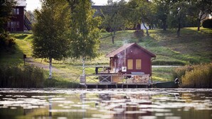 Una cabaña junto al lago con un muelle, rodeada de árboles y una colina cubierta de césped.