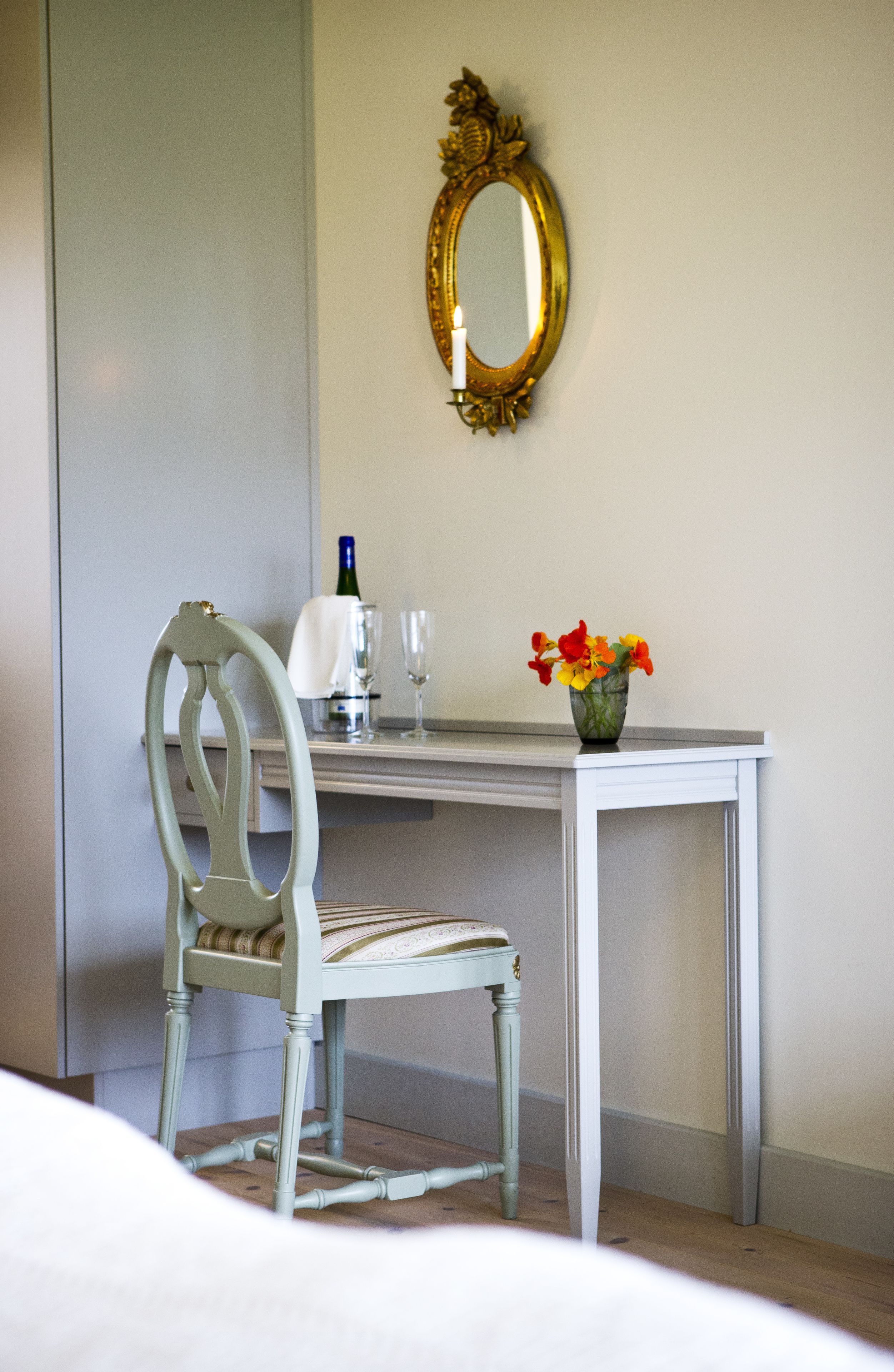 A hotel room with a white desk, a chair, a mirror, a vase with flowers, a bottle of wine, and two glasses.