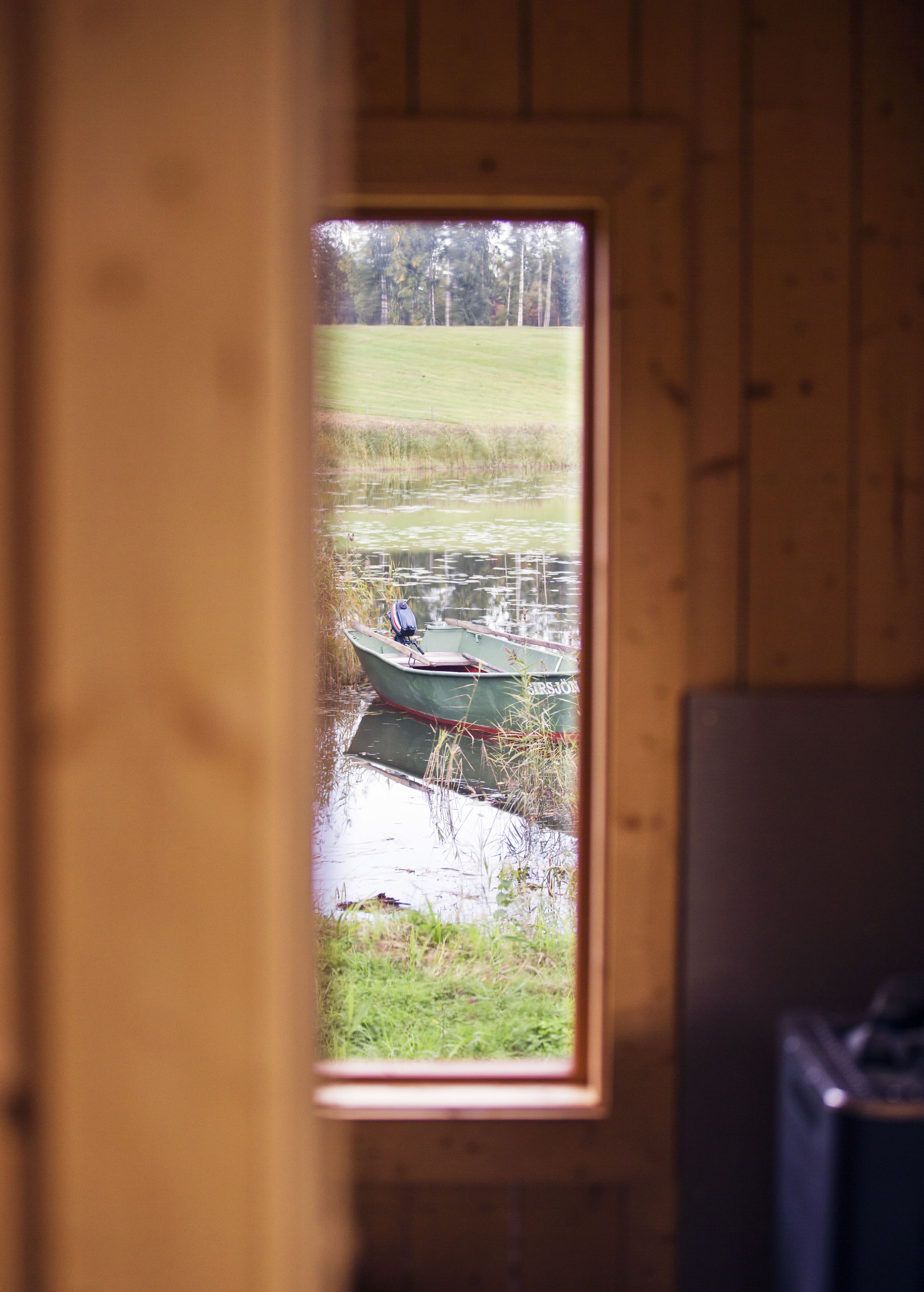 A view through a window of a wooden cabin with a small green boat on a pond.