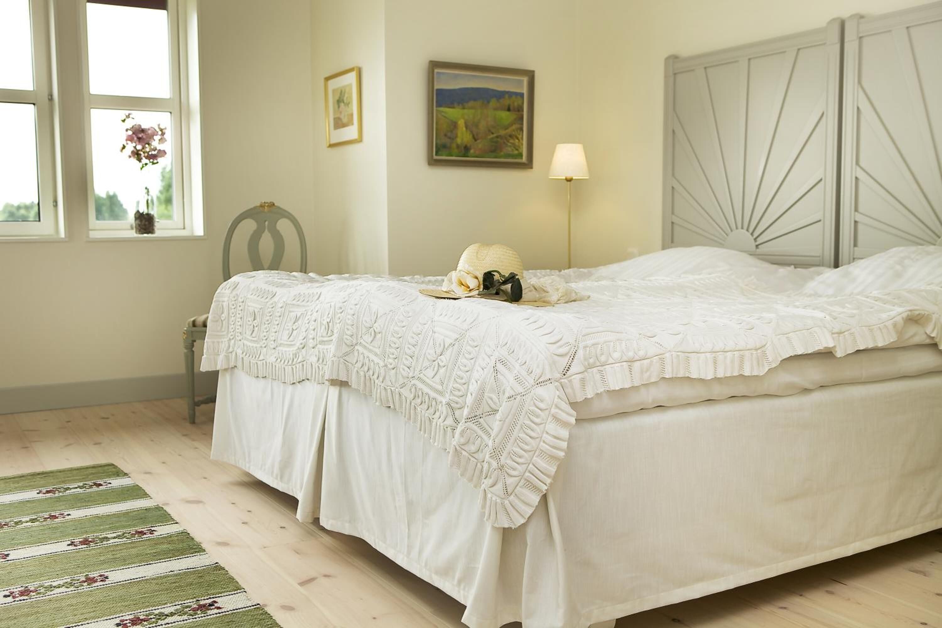A neatly made bed with a white bedspread and lace trim, a bedside lamp, and a framed landscape painting on the wall.