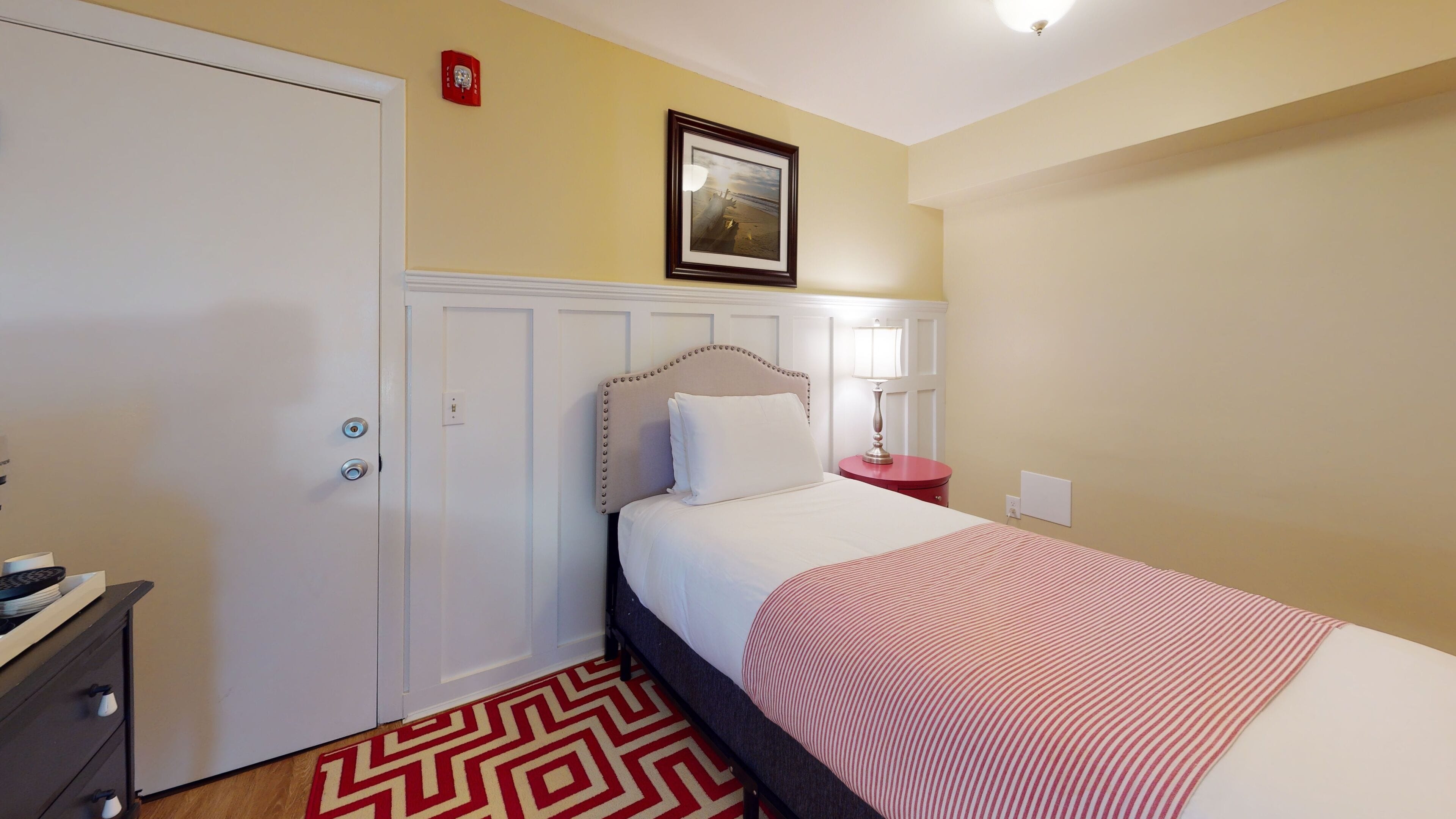 A hotel room with a bed, a nightstand, a lamp, a framed picture on the wall, and a door with a red emergency light.