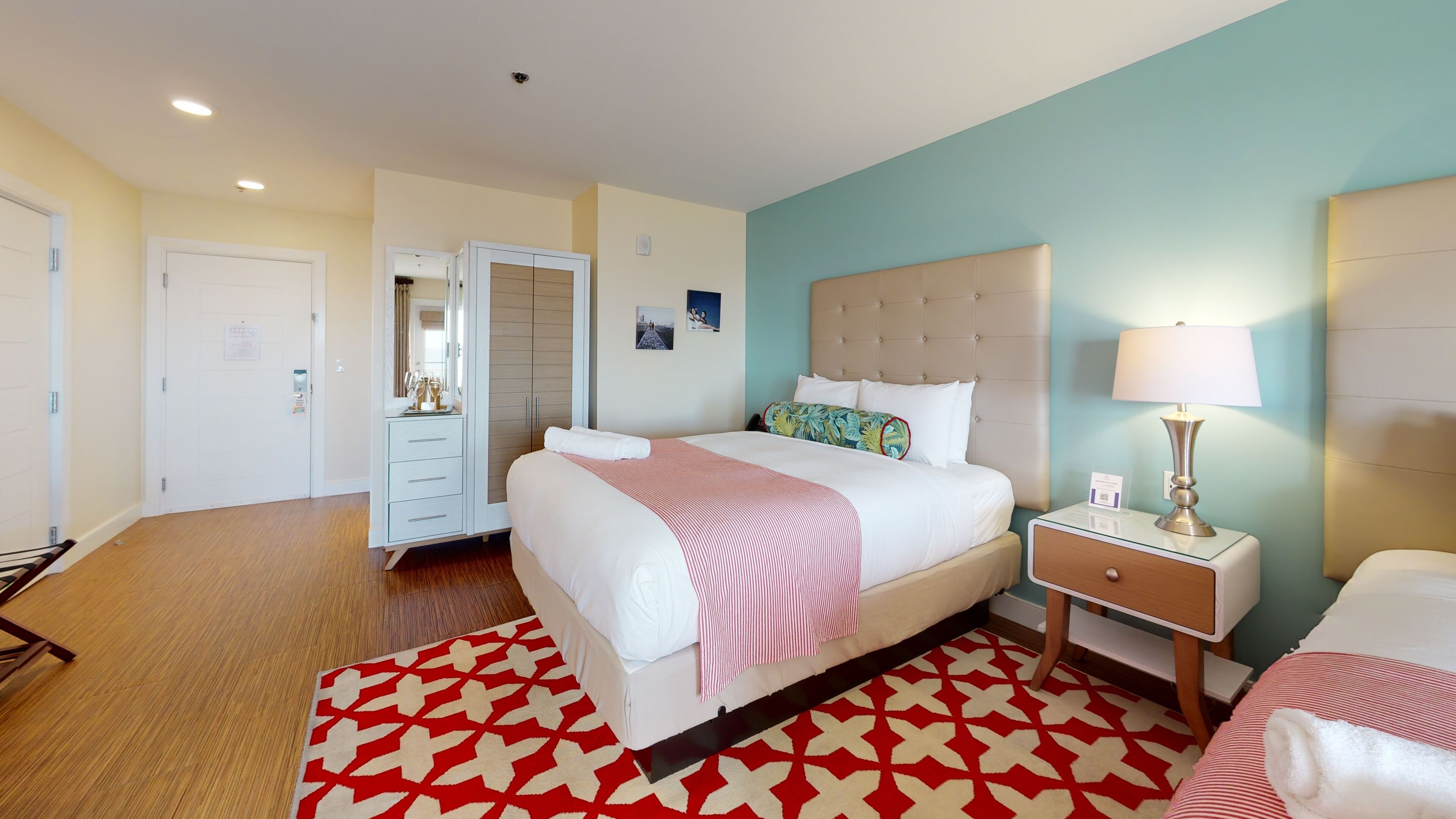Une chambre d’hôtel avec un lit, une table de chevet, une lampe et un tapis aux motifs rouges et blancs.