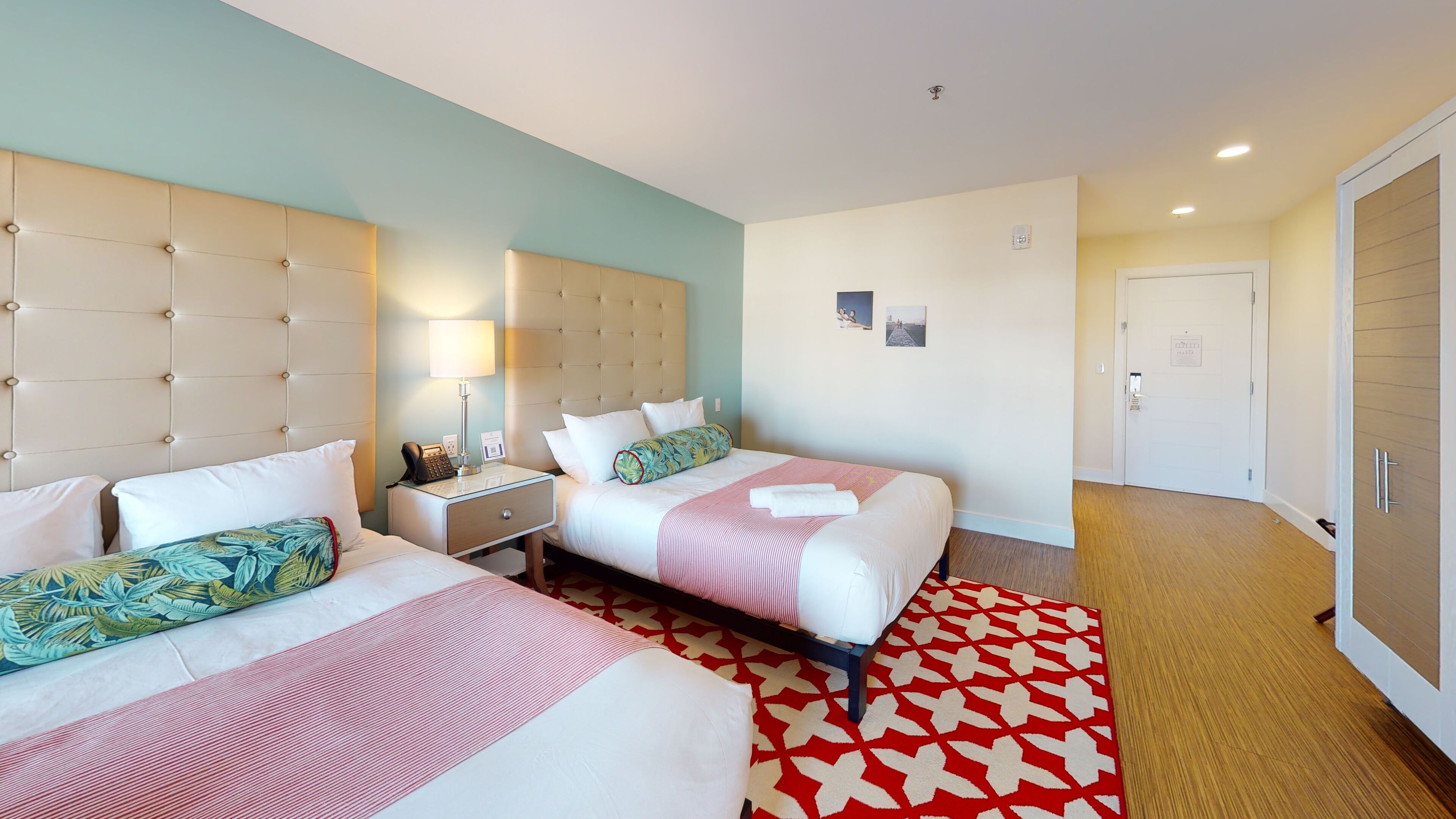 Una camera d'albergo con due letti, un comodino con una lampada, un tappeto rosso e bianco e una porta con un cartello.