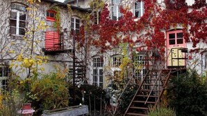 Un patio con una escalera que lleva a un edificio con varias ventanas, algunas con contraventanas rojas, rodeado de plantas y árboles con hojas de otoño.