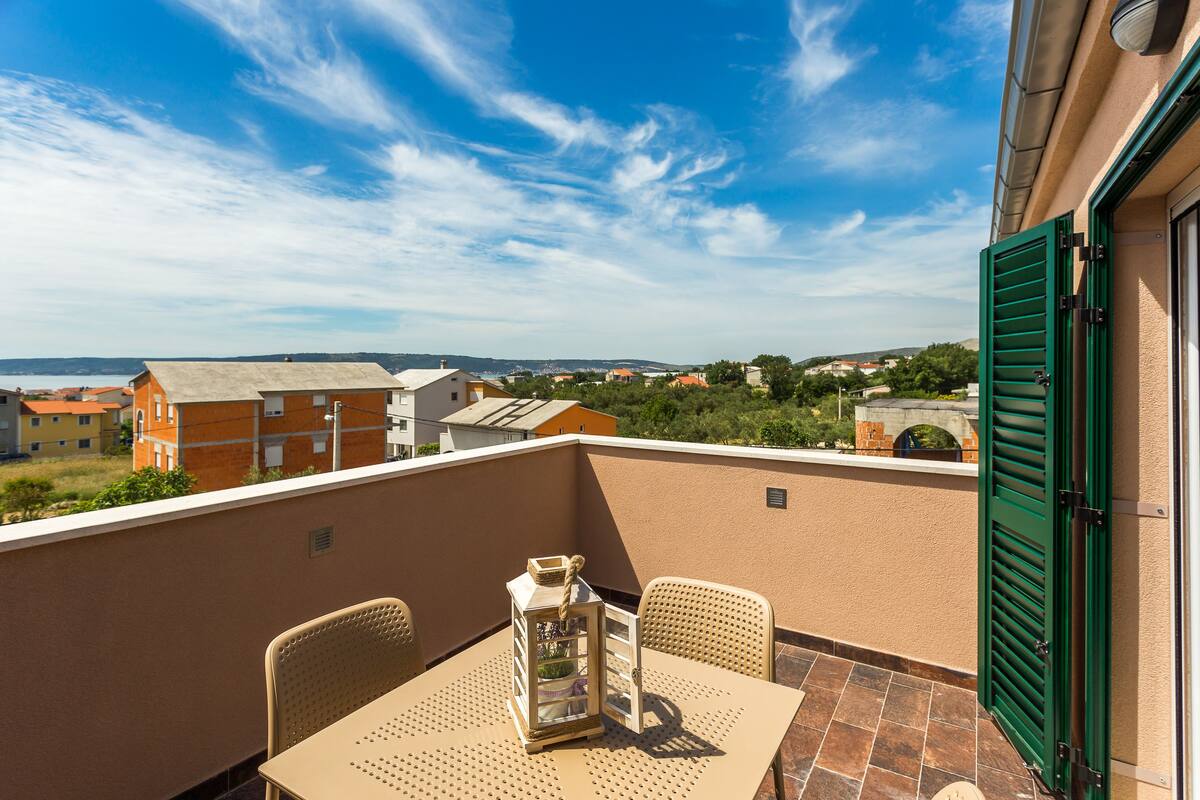 A balcony with a table and chairs, a lantern, and a view of buildings and greenery.