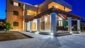 A modern two-story house with a covered patio area, a small garden, and a well-lit entrance. - Villa Rusula (Kastela)