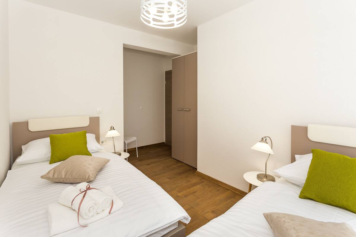 A hotel room with two beds, each with white bedding and green and beige pillows. There is a wooden floor, a small table with a lamp, and a wardrobe.