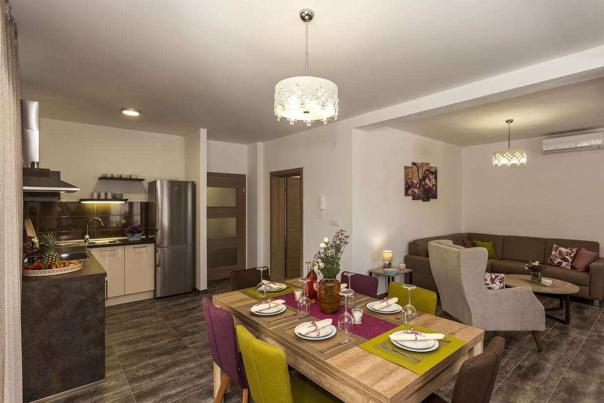 A modern kitchen with a dining area, featuring a wooden table set for six, a sofa, and a chandelier.