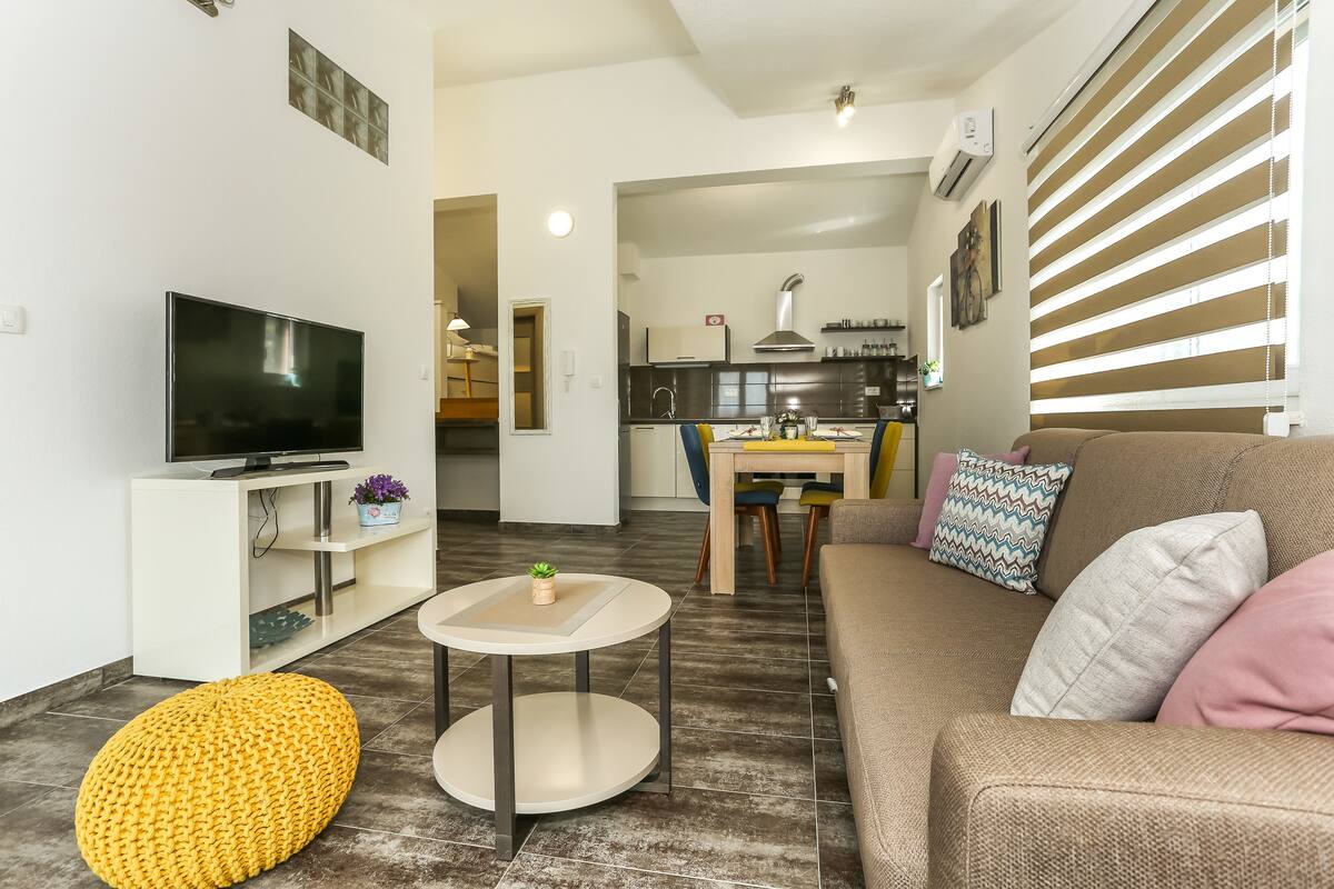 A modern living room with a flat-screen TV, a round coffee table, and a yellow ottoman.