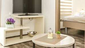 A modern hotel room with a flat-screen TV on a white console, a small table with a candle and a plant, and a bed with a white headboard. - Villa Rusula (Kastela)
