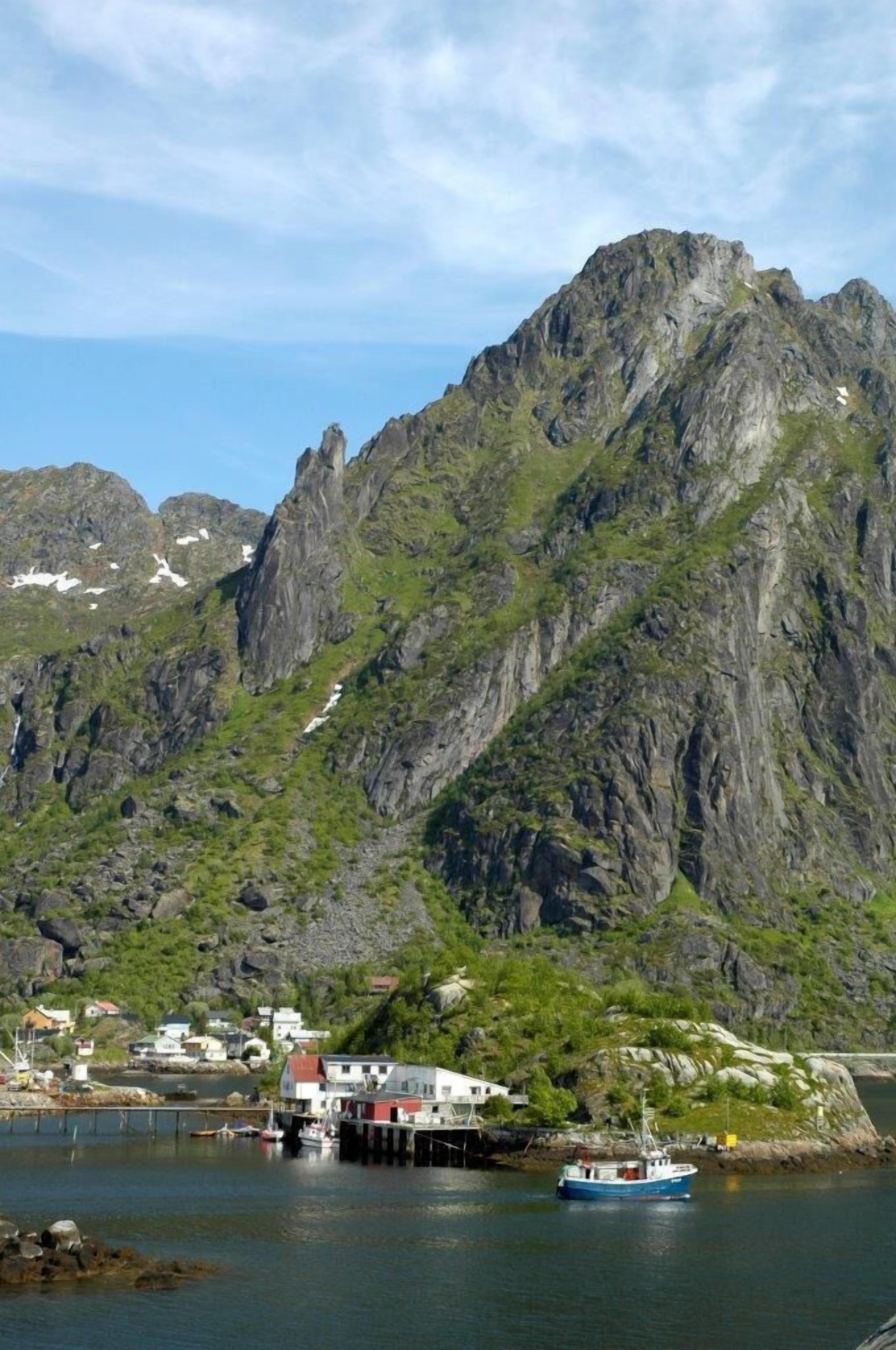 Un village côtier avec une montagne imposante en arrière-plan, une jetée avec un bateau et des maisons le long du littoral.