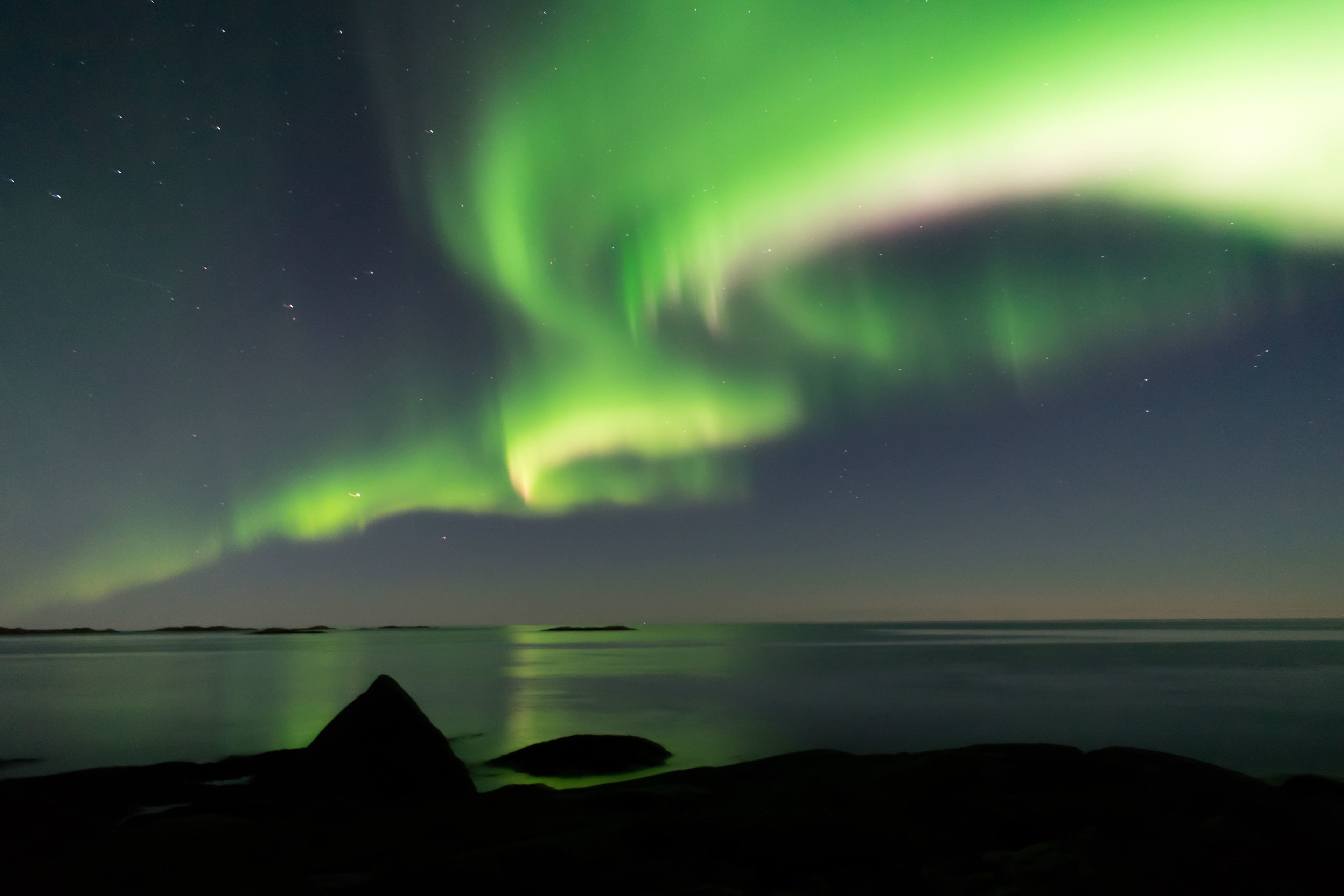 A night sky with the Northern Lights over a calm body of water and rocky shoreline.