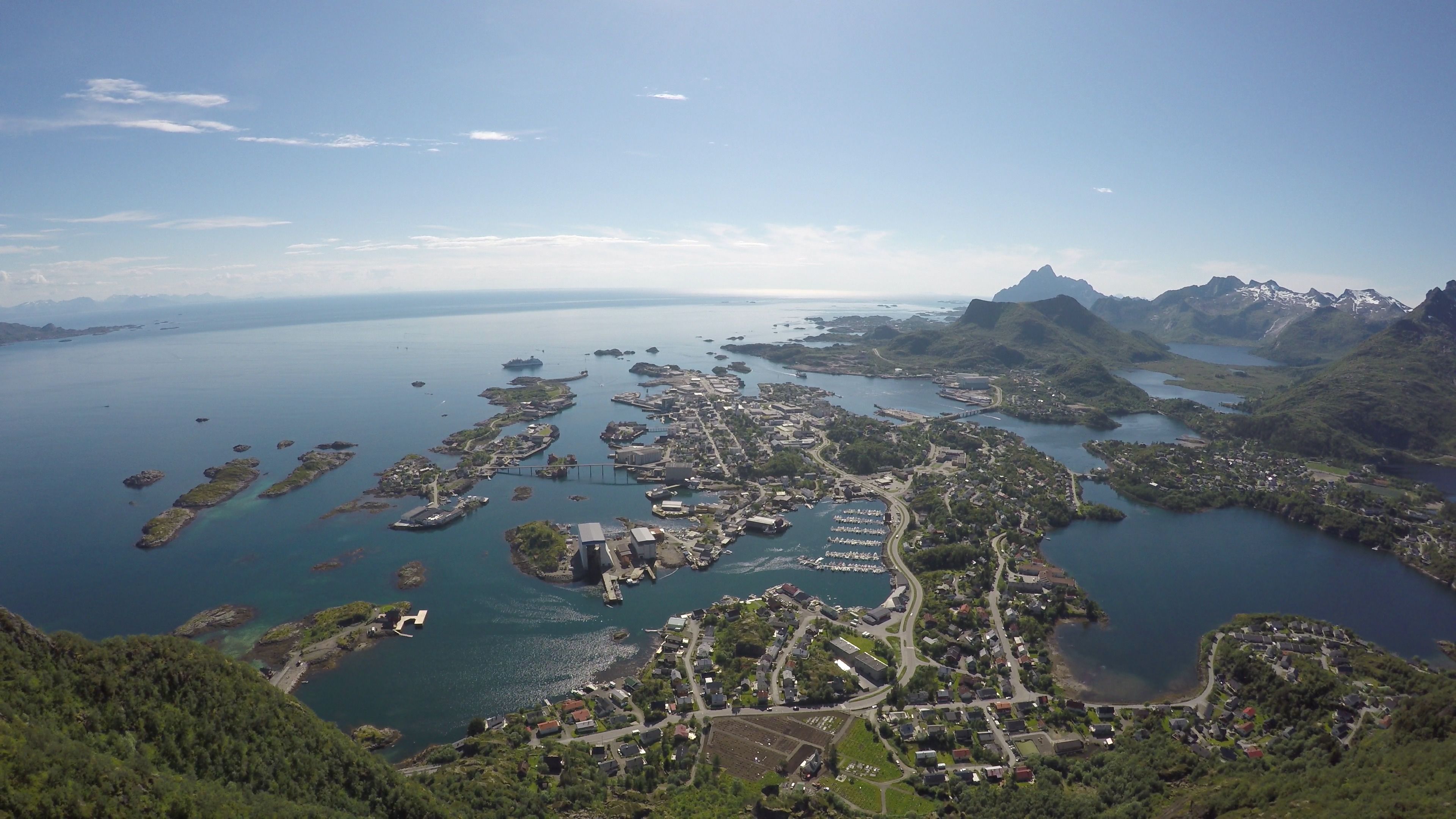 An aerial view of a coastal town with a marina, residential areas, and mountainous terrain in the background.