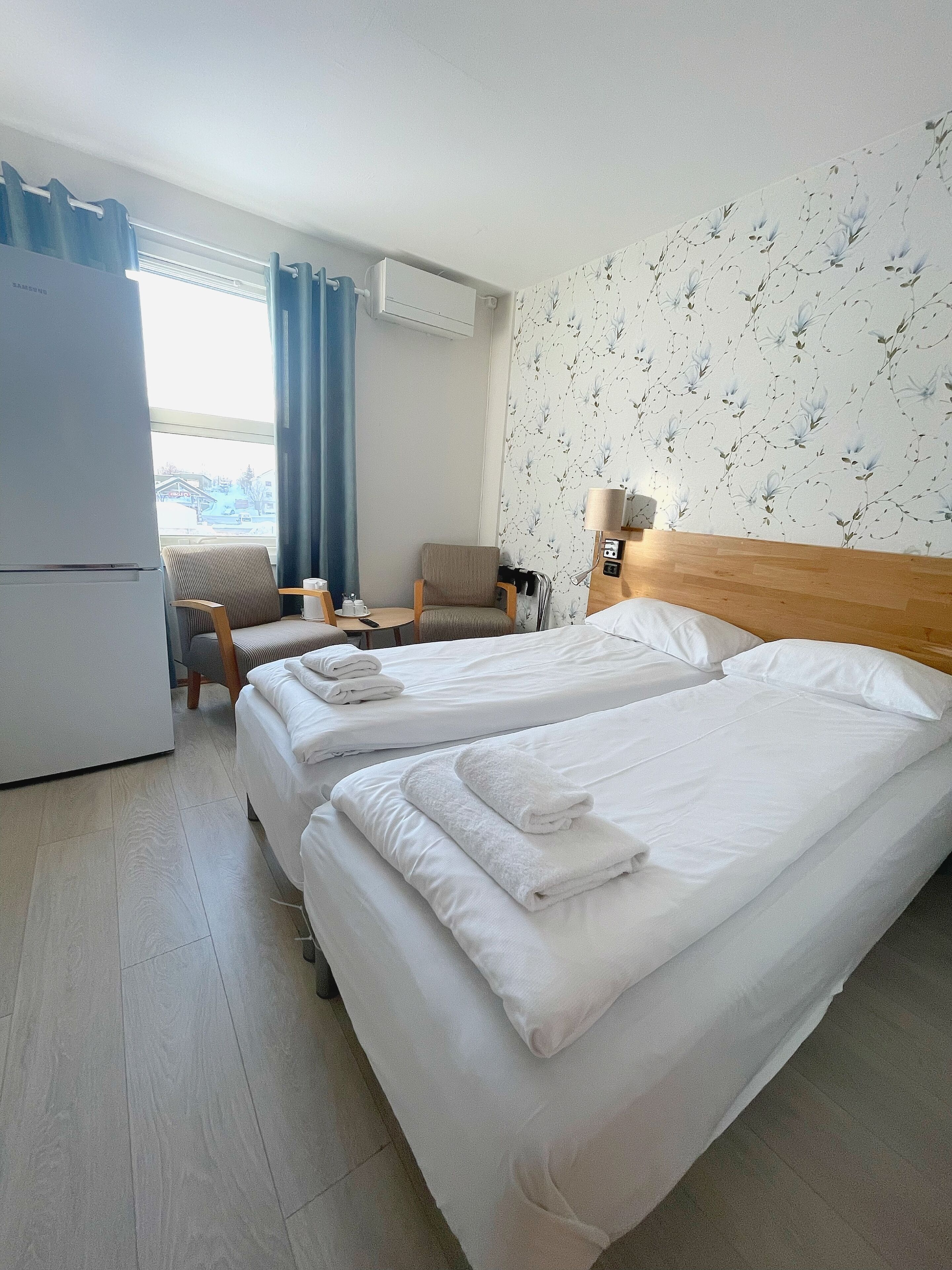 A hotel room with two beds, a small table, a chair, a lamp, and a view of the outside.