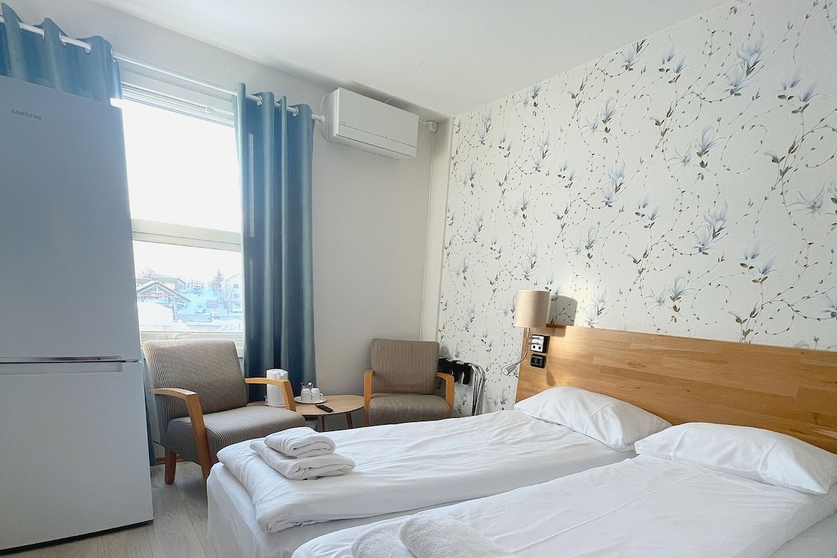 Ein Hotelzimmer mit zwei Betten, einem kleinen Tisch, einem Stuhl, einer Lampe und Blick ins Freie.
