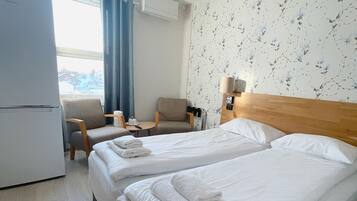 A hotel room with two beds, a small table, a chair, a lamp, and a view of the outside.