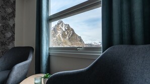 A room with two dark armchairs, a small round table with a candle and a plant, and a window with a view of a mountain. - Marina Hotel Lofoten (Vagan)