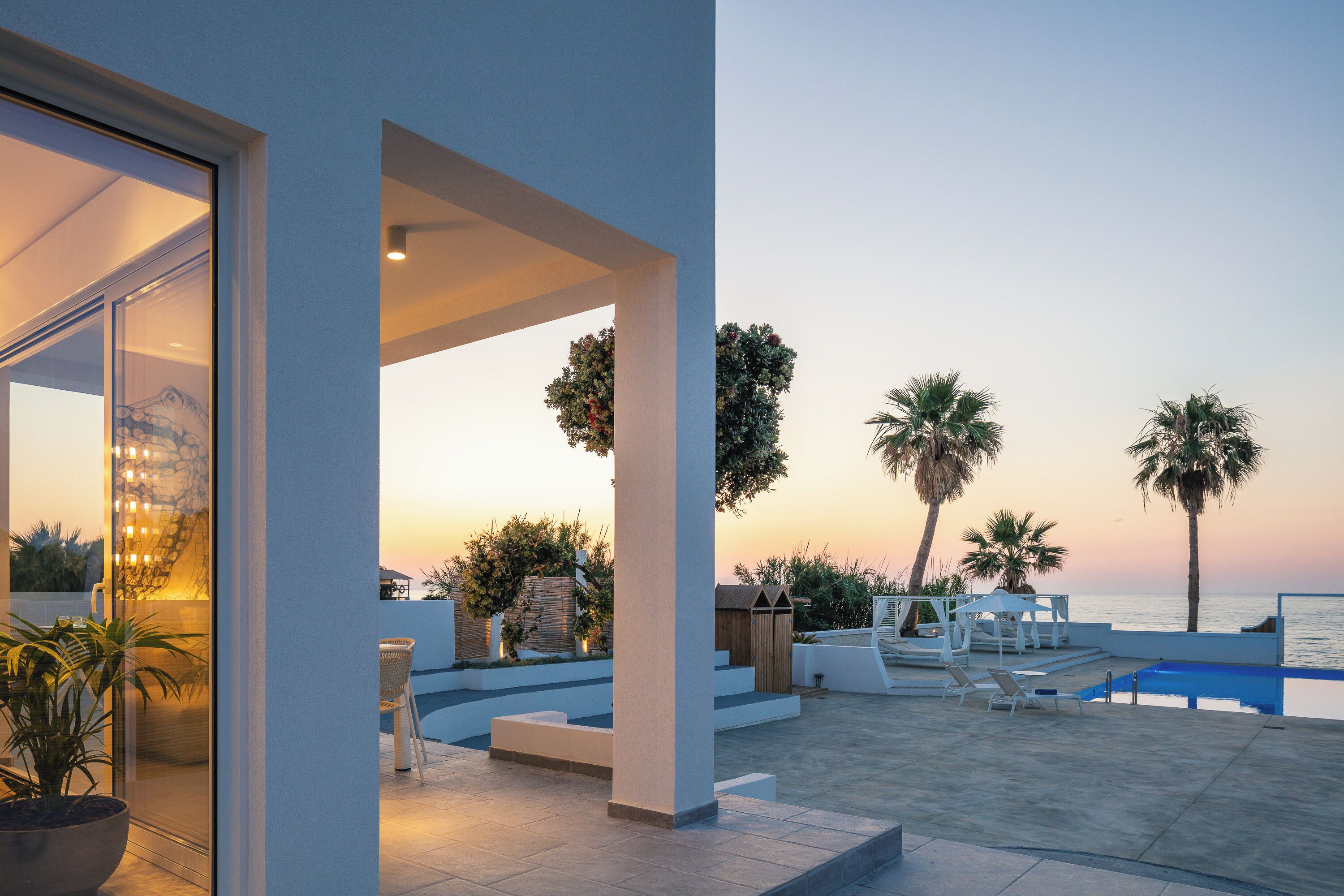 En modern uteplats med pool, solstolar och utsikt över havet vid solnedgången.