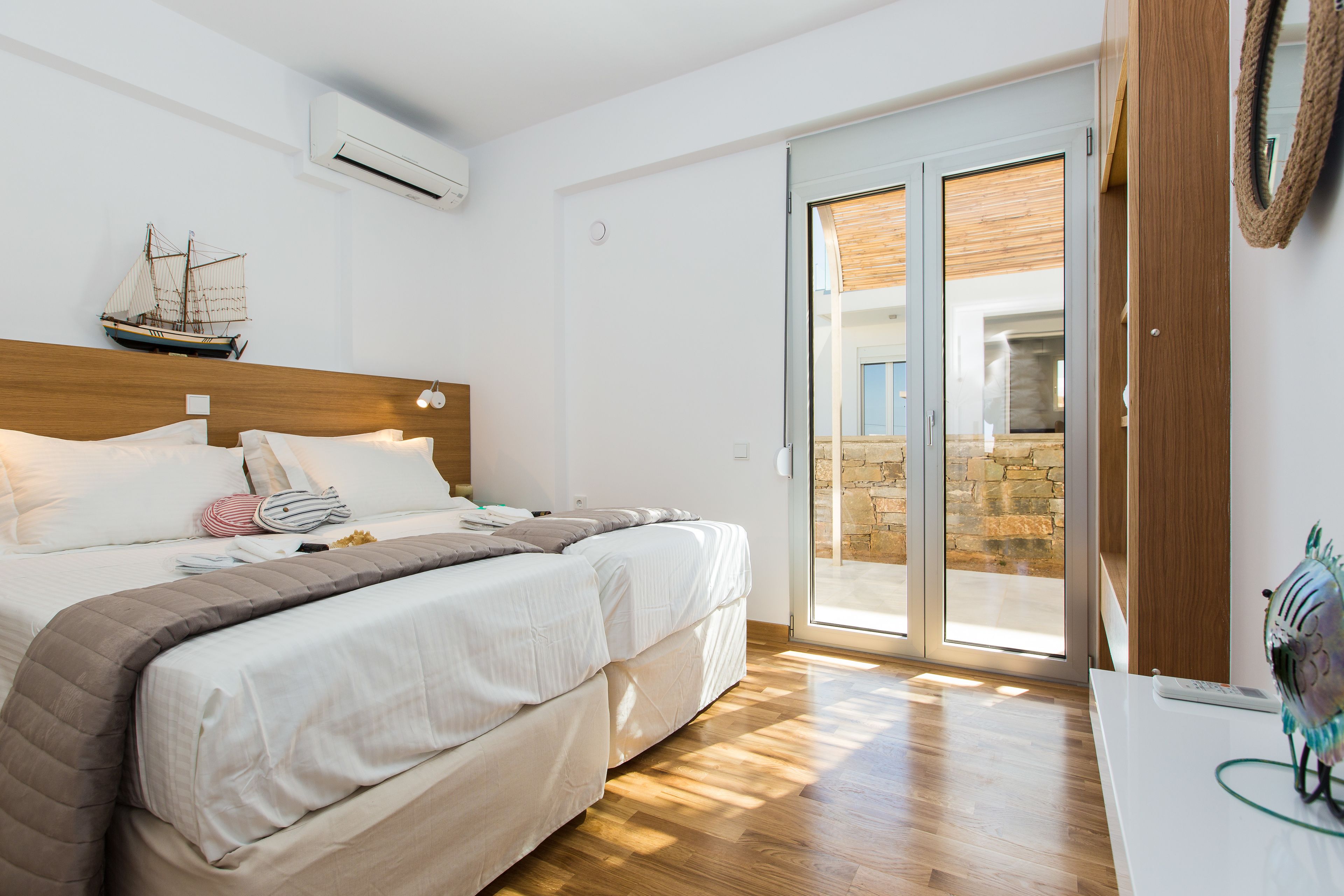 Ein Schlafzimmer mit einem hölzernen Kopfteil, einem Bett mit weißer Bettwäsche, einer Schiffdekoration an der Wand und Blick durch eine Glastür in einen Außenbereich.