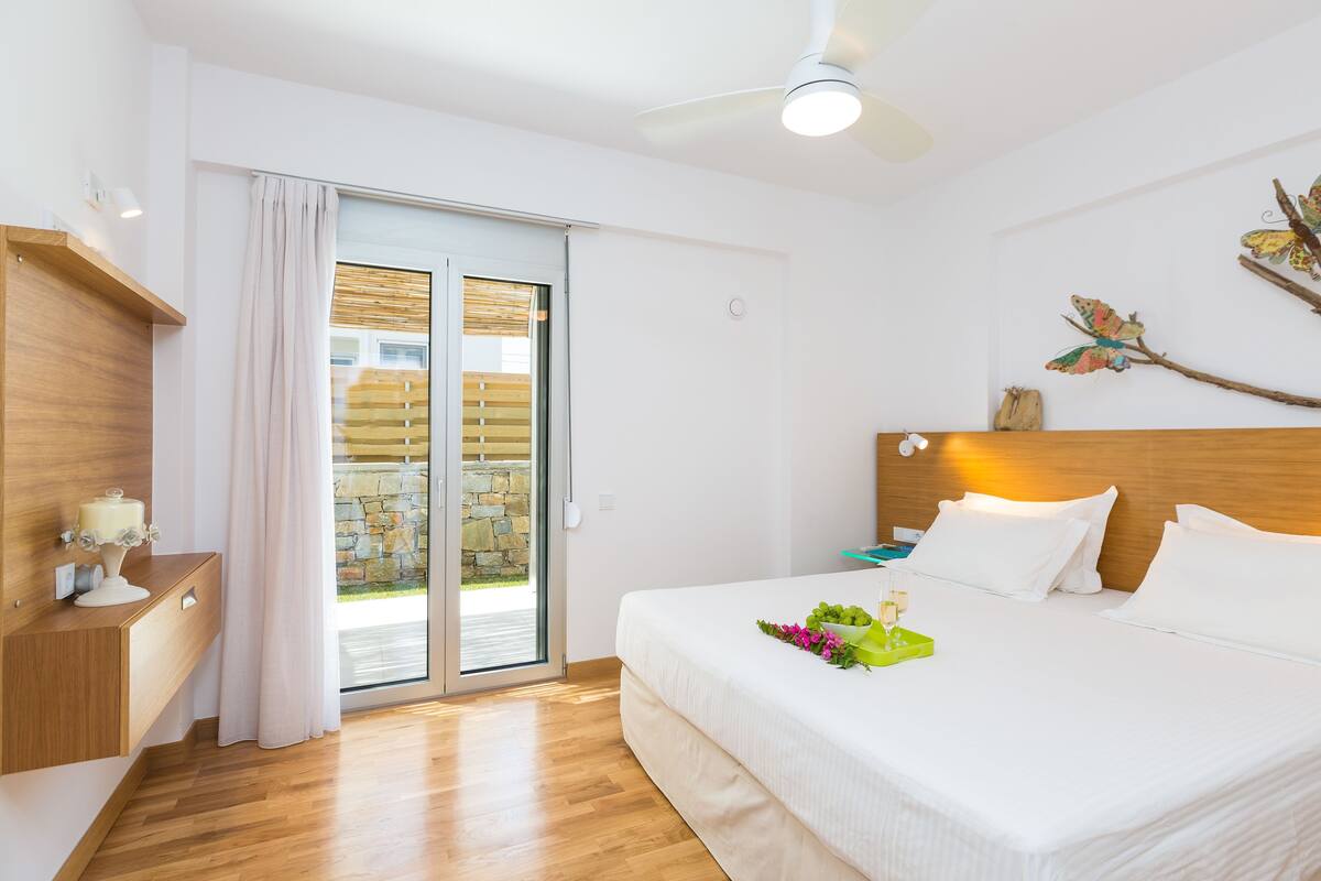 A hotel room with a bed, a wooden nightstand, a ceiling fan, and a view of a stone wall through a glass door.