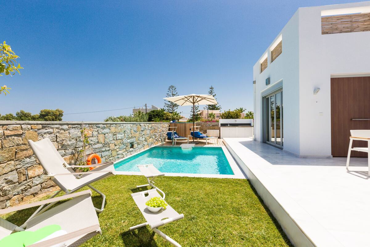 A modern outdoor pool area with lounge chairs, a stone wall, and a clear blue sky.