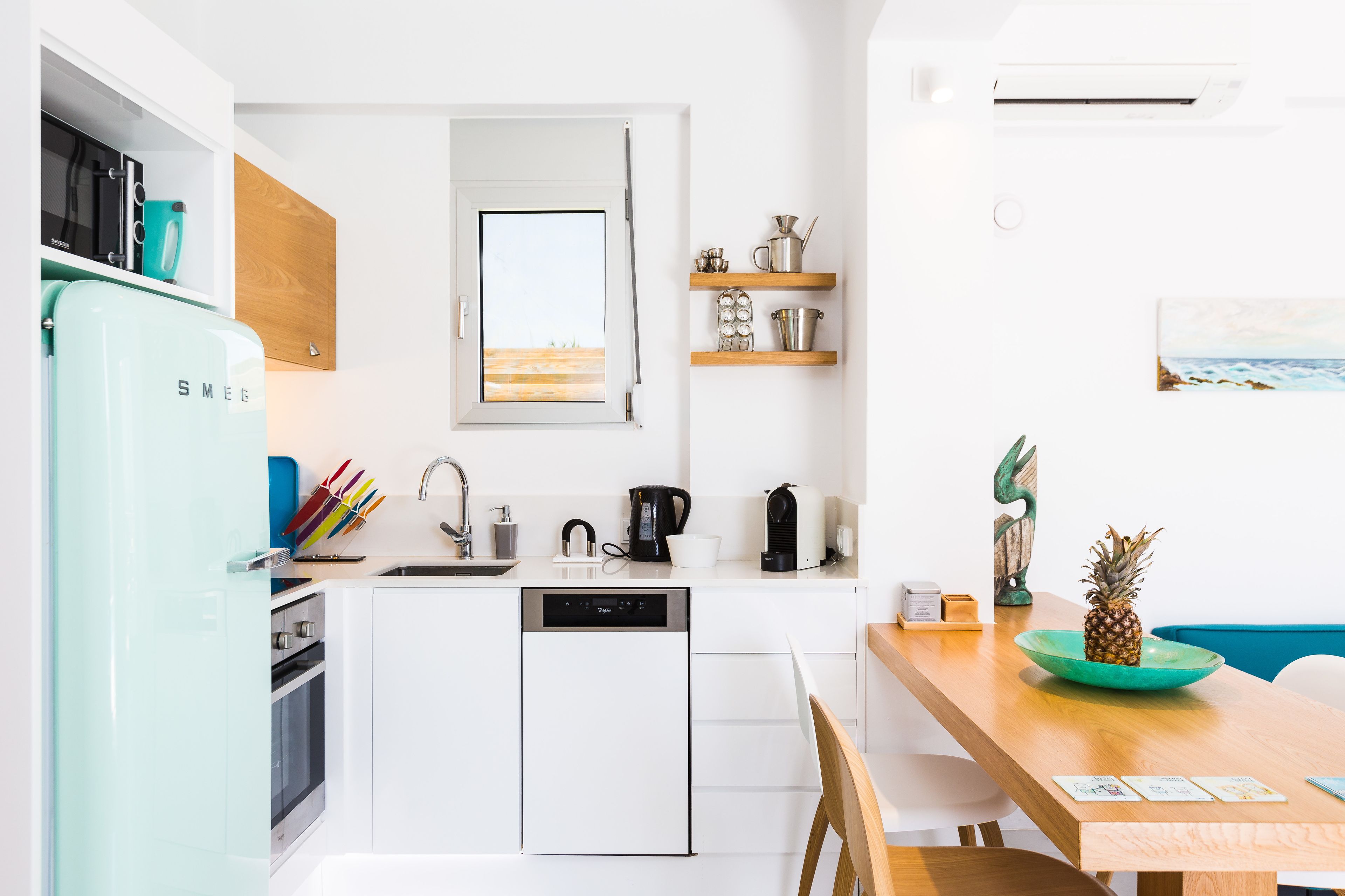 A modern kitchen with white cabinetry, a built-in oven, a dishwasher, and a coffee maker. There is a wooden dining table with a green bowl and a pineapple on it.