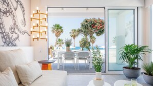 A modern living room with a sofa, a coffee table, and a dining area with chairs and a table. There is a large window with a view of the sea and palm trees. - Thalasses Villas Rethymno (Rethymno)