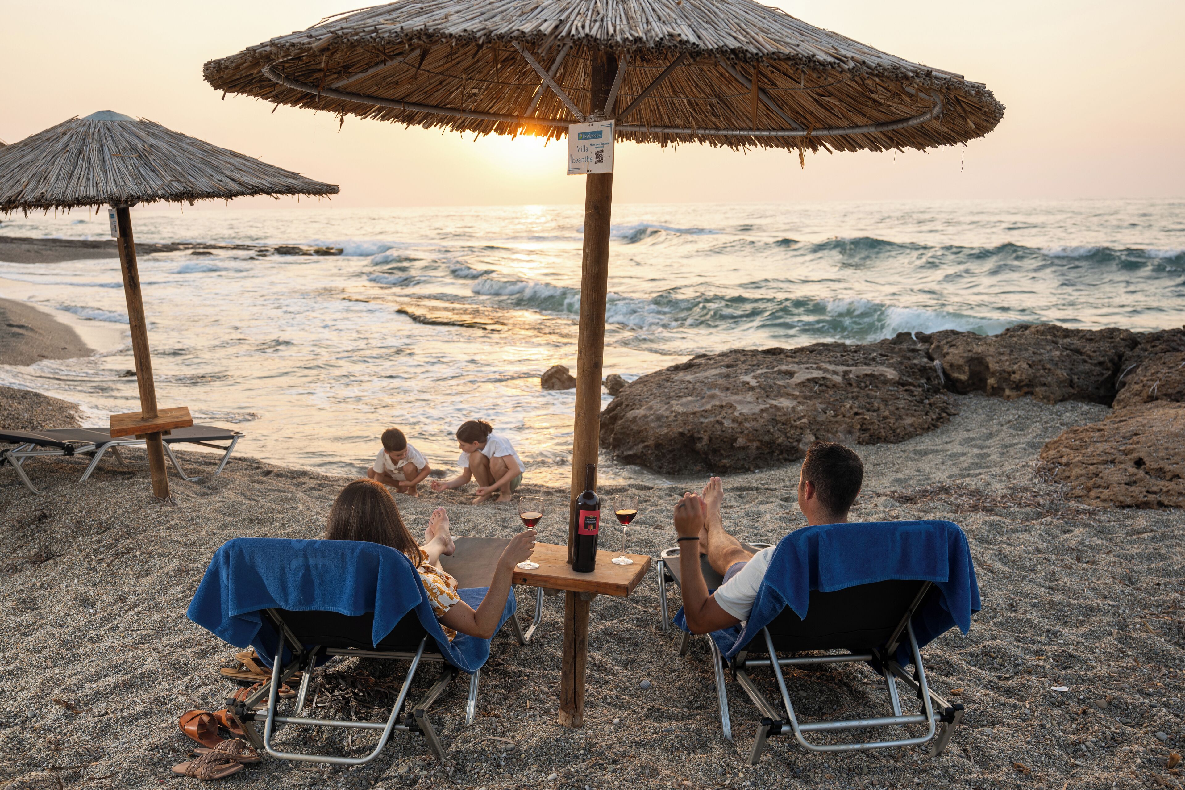 Ein Strandbild mit zwei Liegen unter Sonnenschirmen, einem Tisch mit einer Flasche und Gläsern sowie dem Meer im Hintergrund.