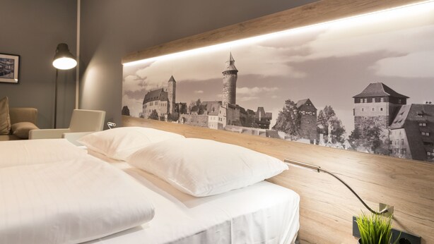 A hotel room with a large bed, a modern desk lamp, and a cityscape mural on the wall. - Aparthotel by dasPaul (Nuremberg)
