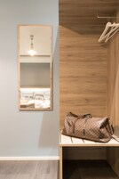 A room with a wooden wall, a hanging light fixture, a mirror with a view of a building, and a bench with a bag.
