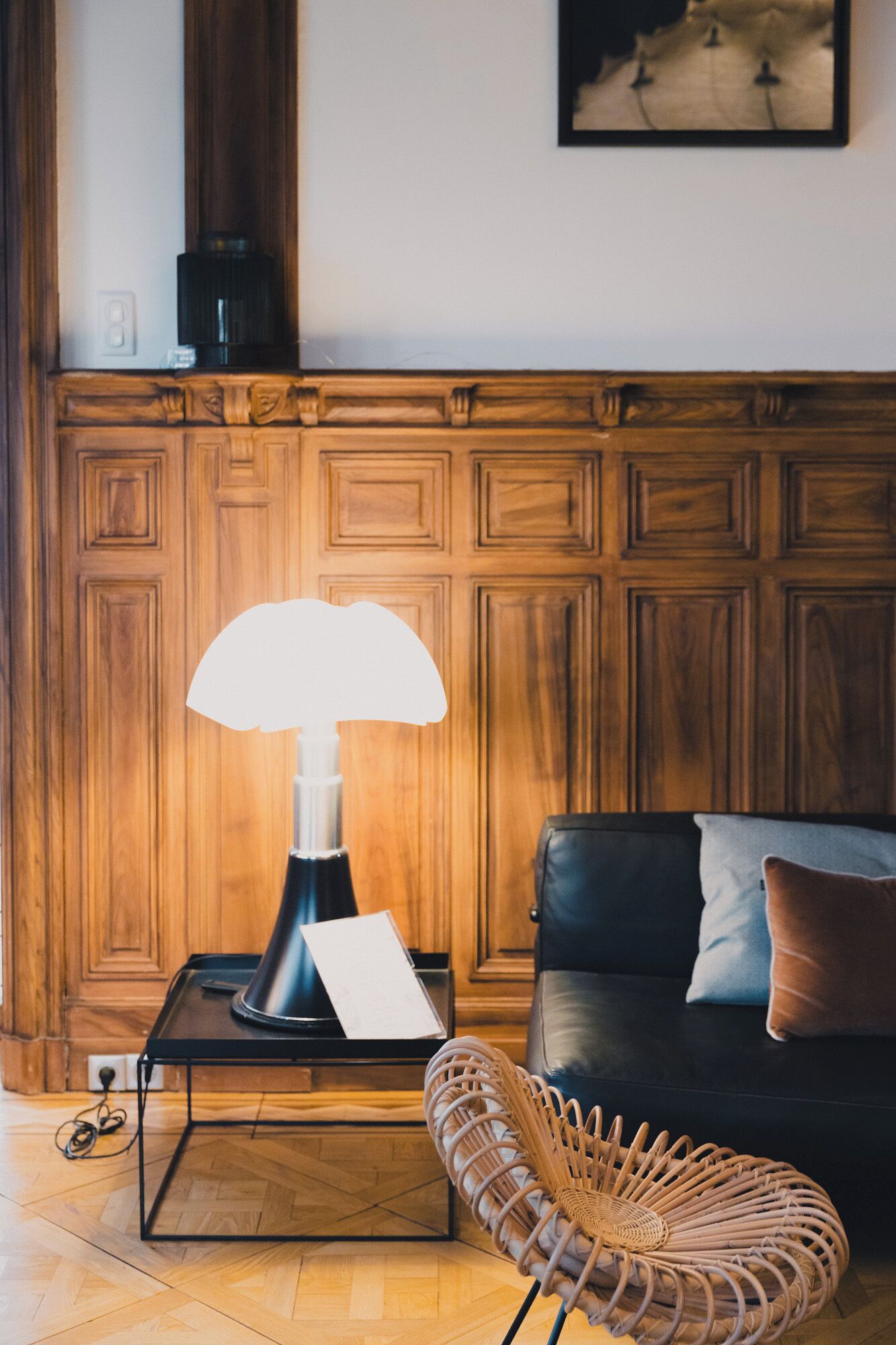 Un salon avec un canapé noir, un fauteuil en osier, une table avec une lampe et un lambris en bois sur le mur.