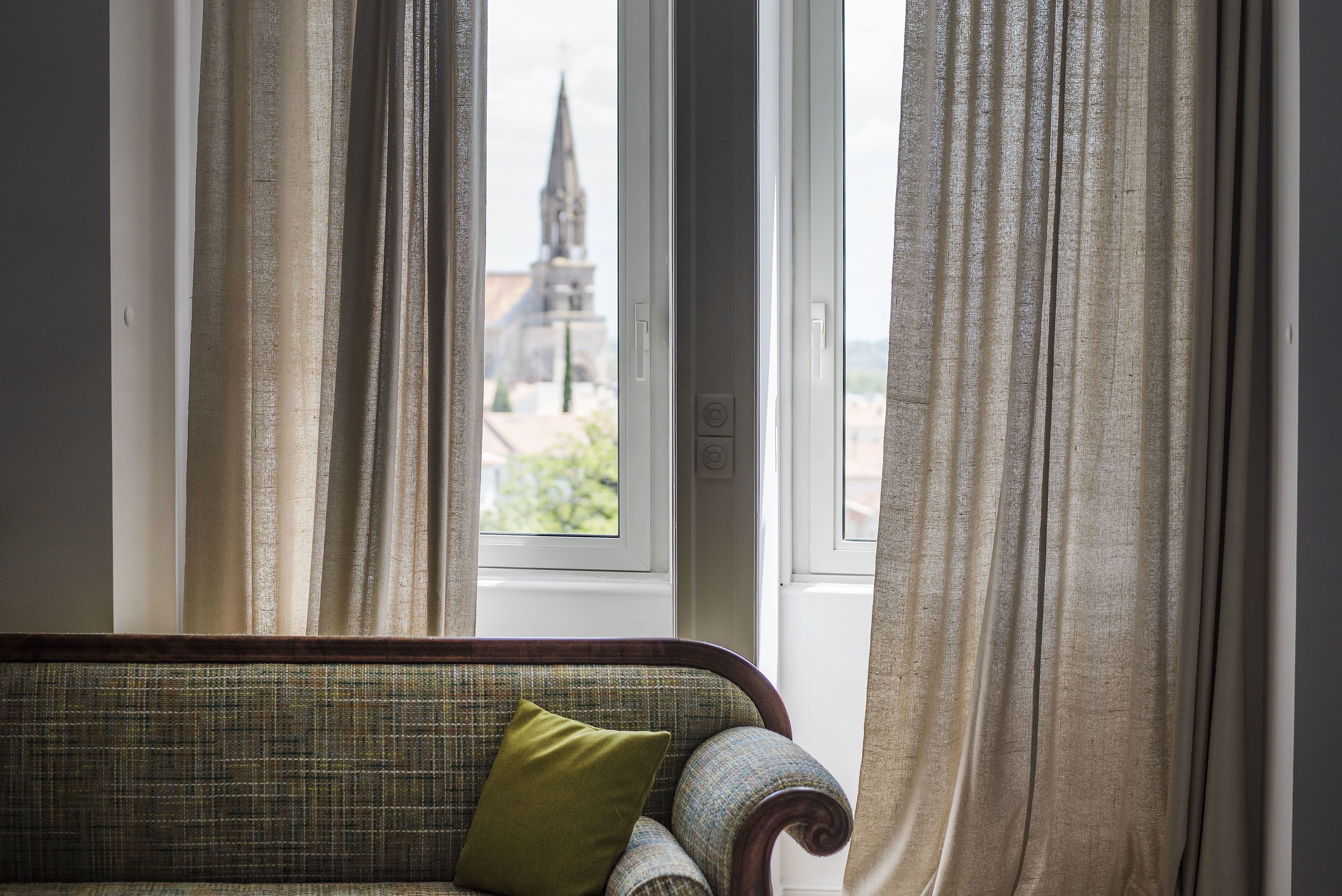 Una habitación con vistas a la aguja de una iglesia a través de una ventana con cortinas.