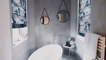 Ein modernes Badezimmer mit einer freistehenden Badewanne, zwei Wandspiegeln und einem kleinen Schminktisch mit Pflegeprodukten.
