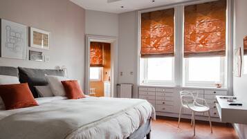 Un dormitorio con una cama grande, un escritorio y una silla. Hay ventanas con persianas anaranjadas, un radiador y cuadros enmarcados en la pared.